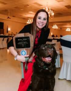 2024 Educational Support Professional of the Year Melissa Wilson stands in front of her school therapy dog holding a trophy.