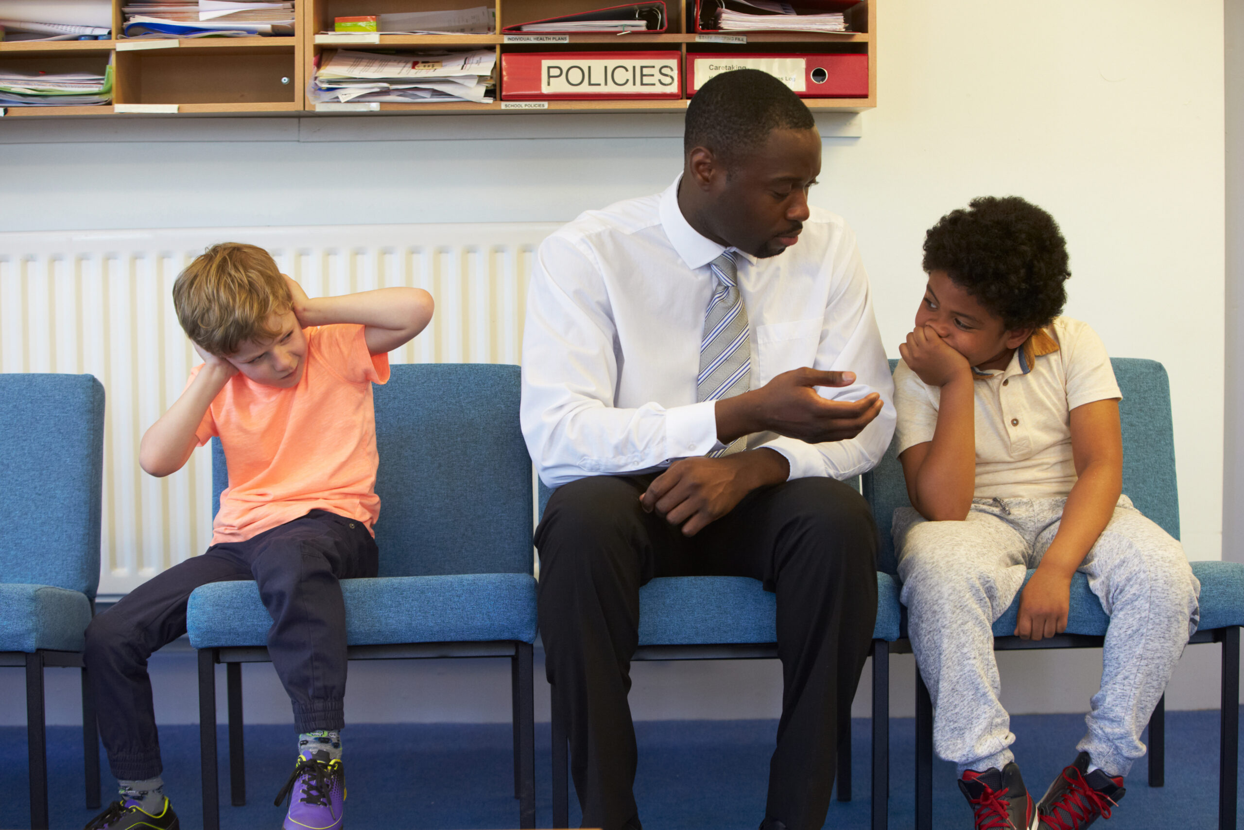 School staff member seated between two elementary-aged students in a school office or counseling setting, speaking with one student while the other sits nearby. Shelves with files and policy binders are visible in the background.